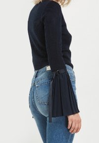 Femme portant un pull noir côtelé avec des manches évasées plissées en forme de cloche et un jean en denim bleu clair, debout devant un fond blanc uni.