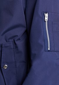 Tmavě modrá bunda z hladké, texturované látky s kapsou na zip a detailem se zapínáním na cvoček na levé straně.