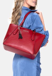 Bolso tote de cuero rojo con dos asas superiores, forma triangular, detalle de borla negra y sutil marca en plata. Textura suave y diseño estructurado.