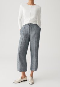 Personne portant un haut à manches longues blanc, un pantalon court bleu ample avec taille à cordon, et des chaussures crème à enfiler, se tenant avec les mains dans les poches.