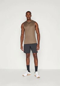 Rjavo brezrokavno športno majico v kombinaciji s črnimi šortami, belimi superge in črnimi nogavicami. Tkanina se zdi lahka in gladka.