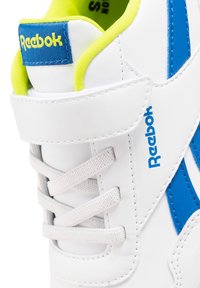 Zapatilla Reebok blanca con logo y franjas azules, cordones blancos, tira de velcro y un forro interior amarillo brillante visible en el cuello del tobillo.