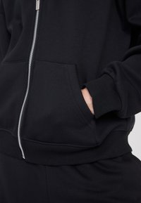 Sudadera con cremallera negra hecha de un tejido suave, presenta un bolsillo frontal, puños y dobladillo acanalados, con detalles de cremallera metálica visibles.