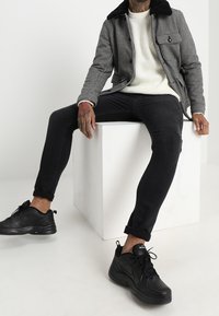 Mustavalkoinen houndstooth-takki mustalla kauluksella, kerma-valkoinen neule, mustat farkut ja mustat lenkkarit, istuu valkoisella kuutiolla.