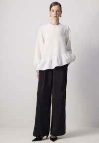 Hvid bluse med lange ærmer og flæse forneden, parret med sorte brede bukser. Stofoverfladen ser tekstureret ud, og outfittet har spidse sorte hæle.