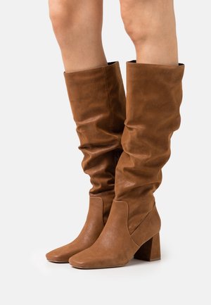 Bottes en cuir marron montantes jusqu'aux genoux avec des talons blocs, portées sur des jambes nues sur un fond blanc.