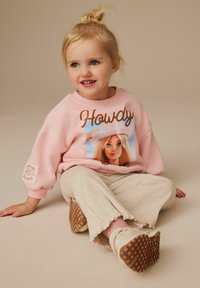 Ljust rosa sweatshirt med texten "Howdy" och en Barbie-grafik, tillsammans med krämfärgade ribbade byxor och vita sneakers med strukturerade sulor.
