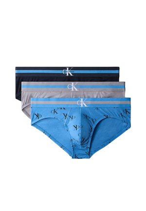 Hombre atlético de piel oscura está descalzo sobre fondo gris, vistiendo calzoncillos negros con cinturilla azul y blanca, sosteniendo los lados de los calzoncillos.