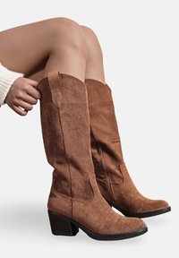 Bottes en daim marron hautes jusqu'au genou avec un bout carré et un talon carré. Présente un accent embossé et une fermeture éclair sur le côté pour un enfilage facile.