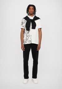 Balta krekls ar melnu greznu rakstu kreisajā pusē, melnas bikses un melns džemperis, kas uzlikts uz pleciem. Baltas teniskates noslēdz apģērbu.