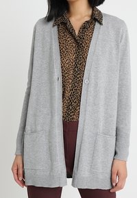 Kvinna i en grå öppen front cardigan över en brun leopardmönstrad skjorta med krage och vinröda byxor, stående mot en neutral bakgrund.