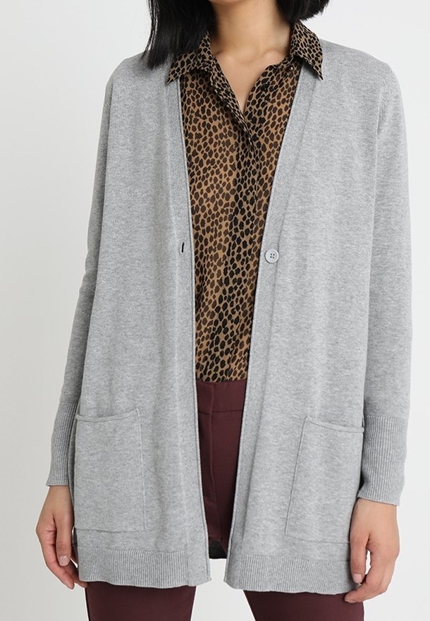Kvinna i en grå öppen front cardigan över en brun leopardmönstrad skjorta med krage och vinröda byxor, stående mot en neutral bakgrund.