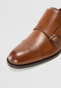 Chaussure habillée en cuir marron avec un bout arrondi, un design à double bride, un détail de boucle en argent et des coutures visibles sur une semelle noire.