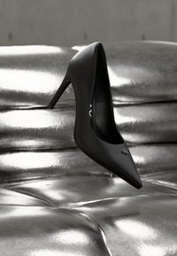 Μαύρο γυναικείο παπούτσι με ψηλό τακούνι πάνω σε ασημένιο μεταλλικό μαξιλάρι με κουμπιά, που δείχνει μυτερή μύτη και λεπτό τακούνι.