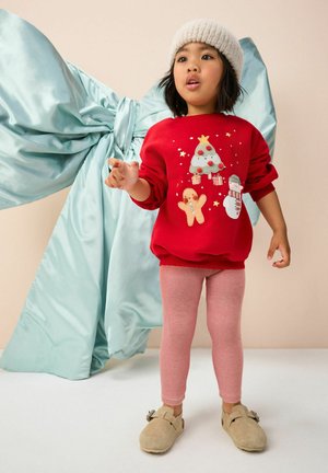 Roter Pullover mit festlichen Grafiken, kombiniert mit rosa gestreiften Leggings und beigen Hausschuhen. Der Hintergrund zeigt einen großen blauen Bogen.