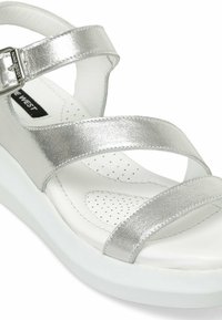 Sandales métalliques argentées avec deux brides croisées, boucle ajustable et semelle blanche rembourrée avec des détails perforés.