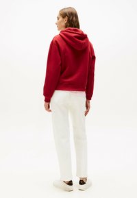 Rode hoodie met een relaxed fit, geribde mouwen en onderuit, gecombineerd met witte rechte broek en witte sneakers met bruine accenten.
