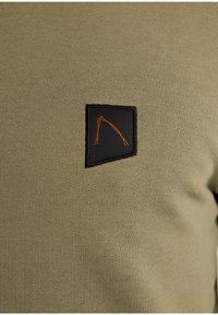 Suéter pullover de color verde oliva con textura de canalé, que presenta un parche rectangular negro con detalles de costura naranja en un diseño geométrico.