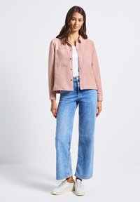 Žena oblečená v bledě růžové košili na knoflíky, bílém topu, světle modrých širokých džínách a bílých teniskách, stojící na jednoduchém pozadí.