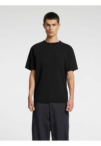 Melna kokvilnas T-krekla ar apaļu kakla izgriezumu un īsām piedurknēm. Brīva piegriezuma ar minimālu detaļu daudzumu, valkāta kopā ar tumšas krāsas biksēm.