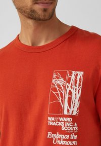 Punane puuvillane T-särk ümmarguse kaelusega, millel on valge puu graafik ja tekst: "WAWARD TRACKS INC. & SCOUTS" ja "Embrace the Unknown."