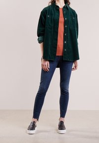 Camicia oversize in velluto a coste verde sopra un top arancione, abbinata a jeans skinny blu scuro e scarpe slip-on nere. Bottoni distintivi sulla camicia.