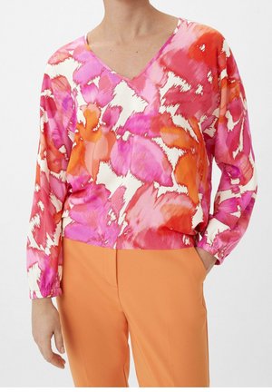 Femme portant un chemisier à manches longues avec un col en V, imprimé floral abstrait rose, rouge, orange et blanc, et un pantalon orange uni.