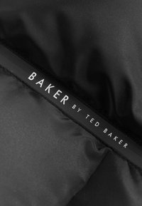Juodas audinys su švelniu paviršiumi, krašte esantis etiketėje užrašyta "BAKER by TED BAKER" baltomis raidėmis.