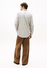 Άνδρας που φοράει ανοιχτό γκρι καρό μακρυμάνικο πουκάμισο και φαρδιά καφέ παντελόνια, στέκεται γυρισμένος προς τα πίσω σε λευκό φόντο.