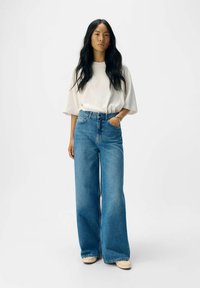 Modre jeans z visokim pasom in sproščenim krojem, ki imajo pet žepov in rahlo zbledel videz. Kombinirani z ohlapno belo majico.