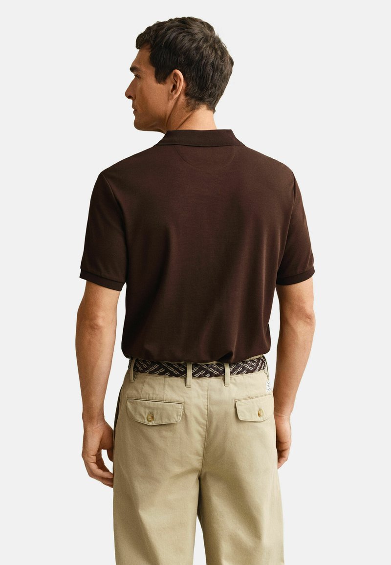 Man draagt een donkerbruin poloshirt met korte mouwen, ingestopt in beige broek met een patroon riem, staand met de rug naar de camera tegen een witte achtergrond.