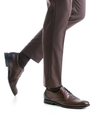 Brauner Lederschuh mit glatter Oberfläche, abgerundeter Spitze und blauen Schnürsenkeln. Socke in dunkler Farbe und eine maßgeschneiderte braune Hose sichtbar.