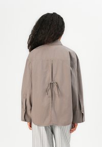 Khaki oversized shirt with long sleeves, back tie detail, relaxed fit, and a center slit. Smooth texture with simple stitching.