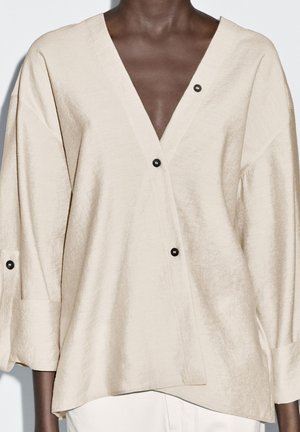 Blouse en lin beige avec un décolleté en V profond, des manches trois-quarts et des accents de boutons noirs. Tissu texturé avec une coupe décontractée.