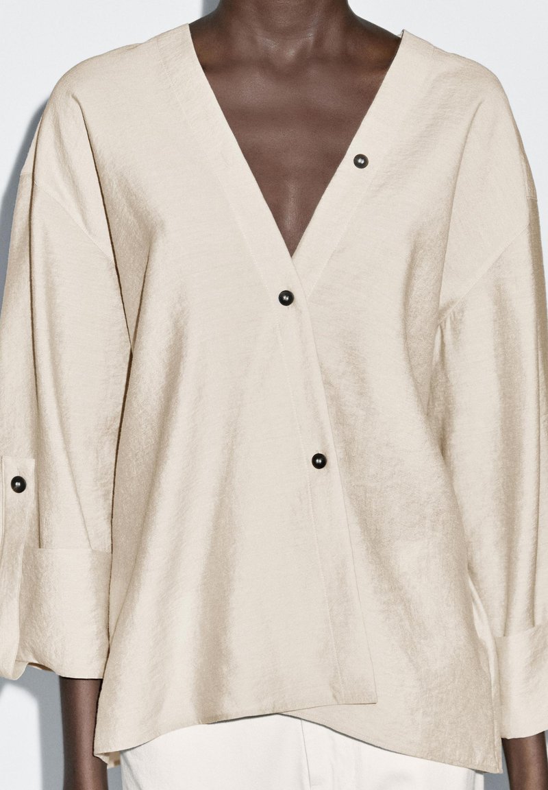 Blusa in lino beige con scollo a V profondo, maniche a tre quarti e dettagli con bottoni neri. Tessuto testurizzato con una vestibilità comoda.