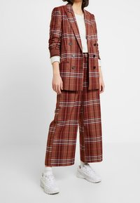 Gepunkteter Anzug in Rot- und Brauntönen mit einem doppelt geknöpften Blazer und weitem Hosenbein. Kombiniert mit weißen Turnschuhen für einen lässigen Look.