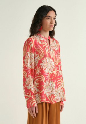 Woman wearing a red floral long-sleeve blouse with a V-neck and brown wide-leg pants, facing sideways against a plain background.