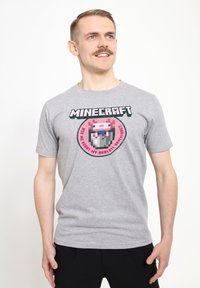 Šedé tričko s krátkým rukávem, které je zdobeno grafickým motivem inspirovaným hrou Minecraft, s růžovými, modrými a černými akcenty v pikselovém stylu v kulatém designu.