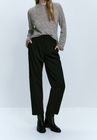 Femme portant un pull gris à manches longues, un pantalon taille haute à jambes larges noir, et des bottines noires, debout les mains dans les poches.
