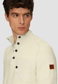 Lichtbeige pullover met een geribde kraag, knoopsluiting, opgestikte zak en een bruine leren logo-accent. Gelaagd stof met zichtbare stiksels.