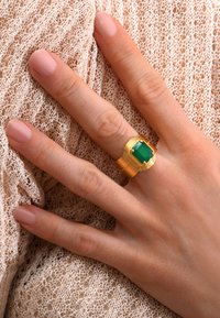 Bague en or avec une pierre verte, présentant des bandes texturées et un sertissage carré. La main repose sur un tissu nude texturé.