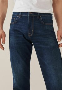 Sötétkék farmernadrág, sportos fazonnal, narancssárga varrással, öt zsebbel és gombos derékmegkötéssel. Sima tapintású.