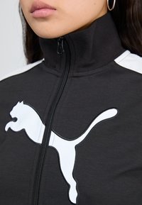Black zip-up jacket with high collar, featuring white Puma logo, contrasting shoulder stripes, and a textured fabric.