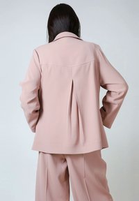 Light pink blazer with a pleated back, notched collar, and long sleeves, paired with matching wide-leg trousers. Smooth fabric texture.