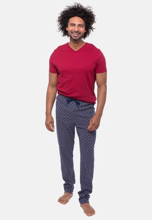 Uśmiechnięty mężczyzna o kręconych włosach, ubrany w czerwoną koszulę z krótkim rękawem oraz granatowe, wzorzyste spodnie od piżamy, stojący boso na białym tle.