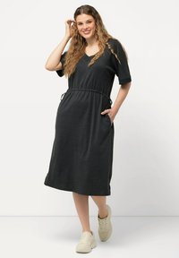 Robe grise foncée à manches courtes, décolleté en V, taille cintrée et poches latérales. Portée avec des baskets de couleur claire. Design simple et décontracté.