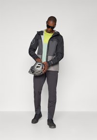 Veste de cyclisme légère noire et grise avec capuche, associée à un t-shirt vert fluo et gris, un pantalon gris et un casque argenté.