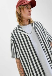 Chemise courte à manches courtes rayée noir et blanc avec un col, des boutons sur le devant et un ourlet arrondi, superposée à un T-shirt gris et une chaîne en argent.