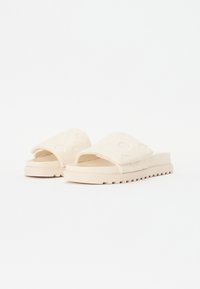 Sandale slip-on bej cu talpă groasă, partea superioară texturată cu modele debrosate subtile și o curea lată peste picior.