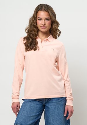 Frau mit welligen braunen Haaren, die ein hellrosa langärmeliges Poloshirt und blaue Jeans aus Denim vor einem schlichten weißen Hintergrund trägt.